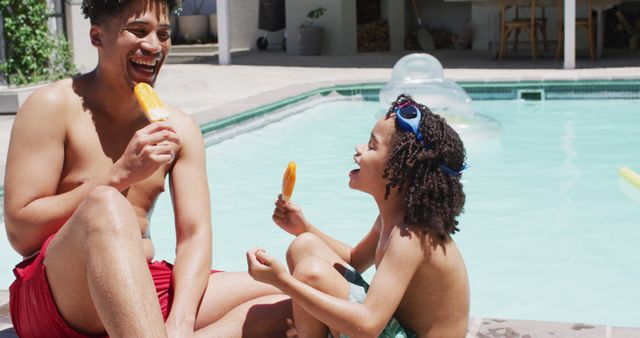 Father and son are sitting by the pool eating popsicles on a sunny day. Both are smiling and relaxed while enjoying their treat. The pool and inflatable toys can be seen in the background. This image captures summer fun and family bonding, ideal for use in advertisements or social media focuses on family activities, summer vacations, or outdoor leisure.