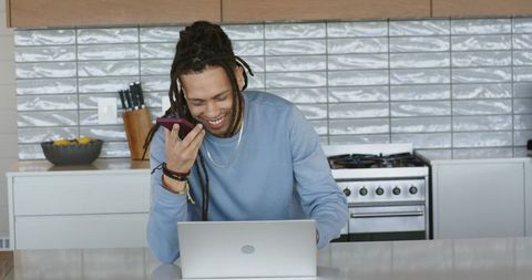 Smiling african american man speaking into smartphone, working on laptop at kitchen island