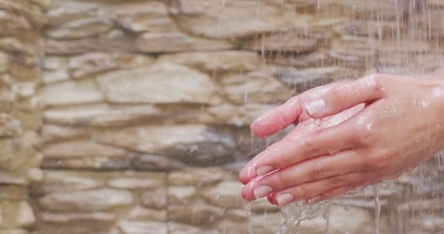 Human hands washing under a stream of running water with a stone background. The focus is on the hands and water, conveying the importance of cleanliness and hand hygiene. This image is ideal for use in campaigns focused on health, hygiene, and skin care products. It can also be used in educational materials about the importance of washing hands to prevent illness.