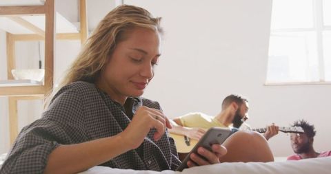 Relaxed Woman Using Smartphone, Friends Playing Guitar in Background
