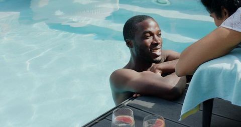 African american man relaxing in pool while indian woman leans on deck with citrus drinks