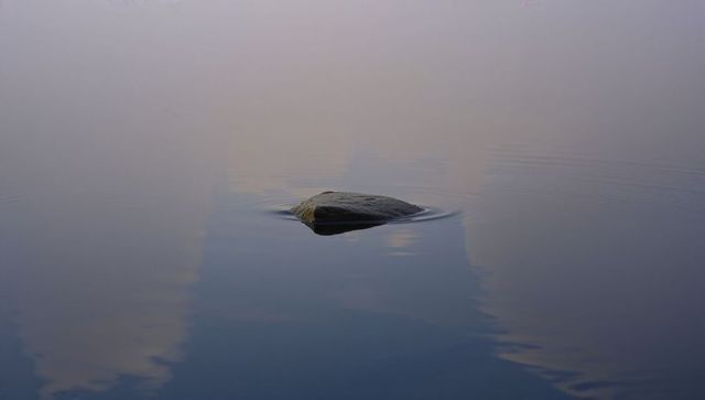 Minimalist solitary rock floating on glassy lake reflecting pastel sky and gentle ripples