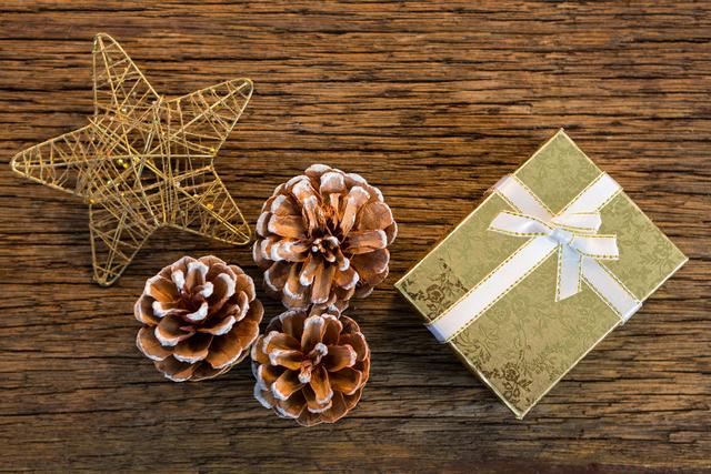 This image features a Christmas gift wrapped in gold paper with a white ribbon, accompanied by pine cones and a decorative star on a wooden background. Ideal for holiday greeting cards, festive invitations, seasonal blog posts, and social media content celebrating Christmas and winter holidays.