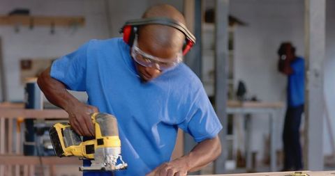 Woodworker operating jigsaw in workshop with colleague nearby