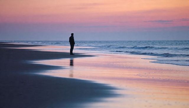 Solitary silhouette standing on pastel shoreline at sunset reflecting on wet sand and sea