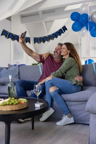 Middle-aged couple celebrating birthday at home taking selfie on cozy sofa
