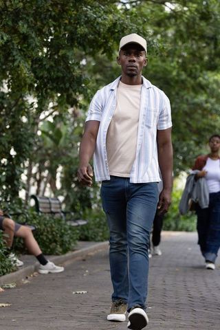 African man walking on cobblestone park path wearing baseball cap, jeans and sneakers