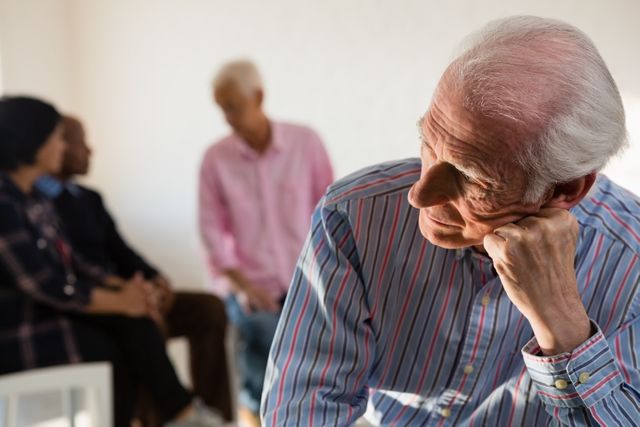 This image depicts a senior man deep in thought with friends in the background at an art class. It is ideal for use in articles or advertisements related to senior activities, retirement communities, mental health, social engagement, and creative hobbies for the elderly.
