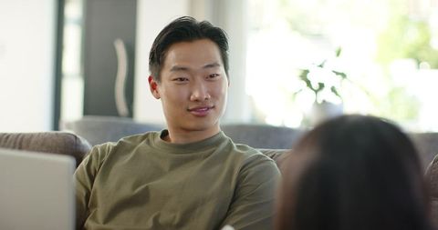 Asian man and partner chatting on gray couch with laptop in bright living room
