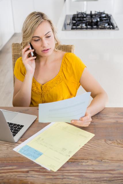 Woman in casual yellow top talking on mobile phone while looking at bill in her kitchen at home. Laptop and various papers on table suggest financial duties or personal expenses. Ideal for illustrating home office, budgeting, financial planning, or remote work themes.