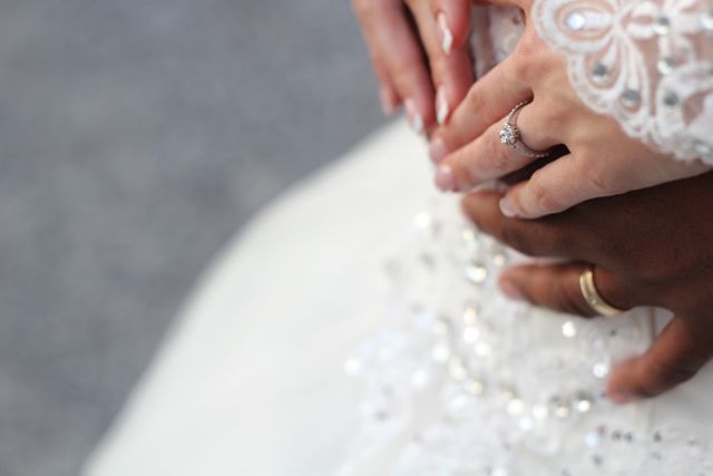 Close-up of bride's and groom's hands tenderly intertwined, both showing wedding rings. Image captures heartfelt emotions and intimacy of wedding day. Useful for wedding invitations, blog posts about marriage, multicultural love, romantic advertisements.