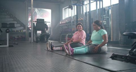 Two women stretching together in gym wearing mint and pink activewear for partner workout