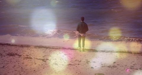Solitary man standing at ocean shore facing waves with dreamy lens flare bokeh atmosphere