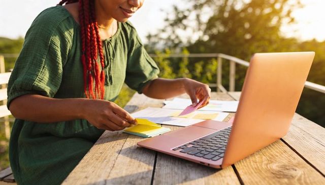 Braided woman organizing sticky notes at outdoor laptop workspace with rose-gold laptop