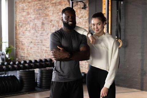Smiling black male trainer and asian client posing in urban gym wearing smartwatches