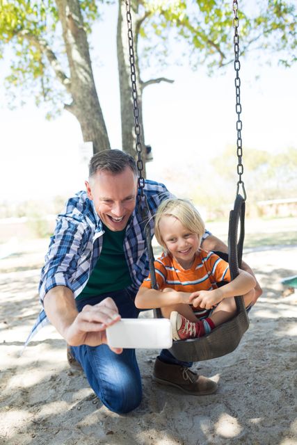 Happy father taking selfie with son sitting on swing at playground