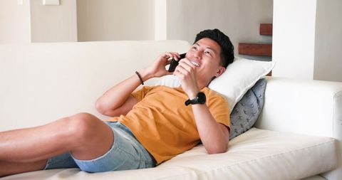 Asian Man Relaxing on Sofa at Home Talking on Smartphone