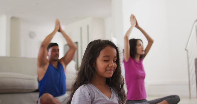 This image shows a peaceful family engaging in yoga and meditation together in their home. The child sits between the mother and father, eyes closed and hands in prayer position, embodying calmness and tranquility. This is perfect for promoting family wellness, mindful living, and healthy lifestyle activities. Ideal for use in health and fitness blogs, websites, or advertising related to family bonding, meditation practices, or holistic health initiatives.