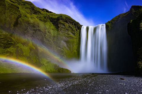 Cascading waterfall with double rainbow over moss-covered cliffs and black pebble shore