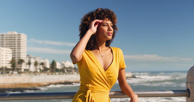 Young woman in a yellow dress enjoying a coastal view on a sunny day. Background shows an urban coastal area with buildings, water, and sky. Ideal for use in lifestyle blogs, travel websites, fashion articles, and summer holiday campaigns.