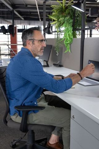 Middle-aged man sitting at desk reaching for laptop in modern open-plan office with greenery