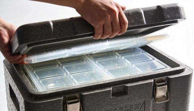 Hands lifting foam lid revealing insulated meal cooler with modular clear food trays