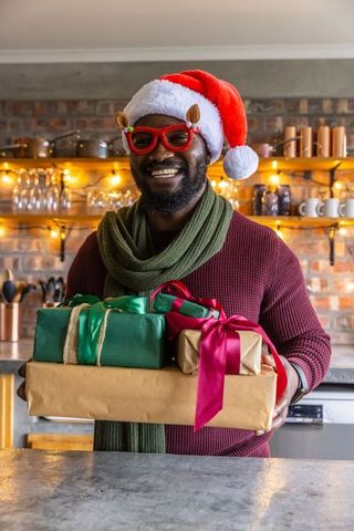 Smiling african american man holding christmas gifts wearing santa hat in cozy kitchen