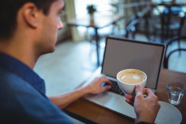 Man holding coffee cup while using laptop in a cafe. Ideal for illustrating remote work, freelance lifestyle, productivity, and modern work environments.