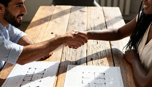 Shaking hands over blueprints on rustic wooden table during daylight business meeting