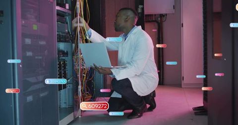 Crouching IT technician inspecting server rack and colorful network cables with laptop