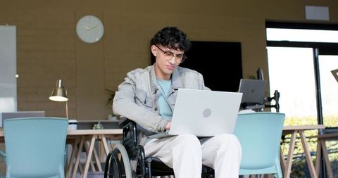 Young man in wheelchair working on laptop in bright coworking space, inclusive workplace