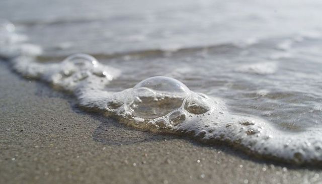 Translucent bubble nestled in seafoam on sandy shoreline, macro ocean close-up