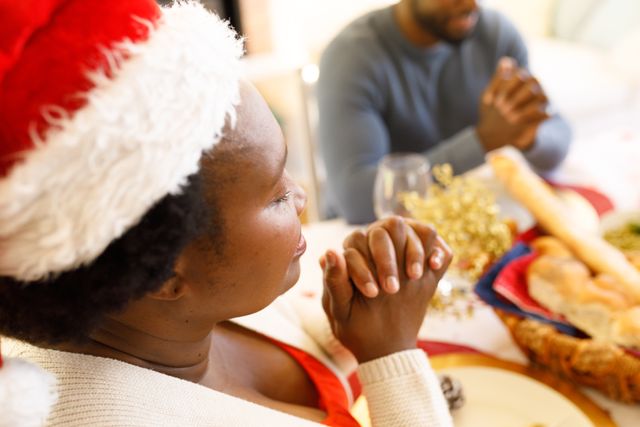 Side view of an african-american woman wearing a santa hat praying with family at a dinner table with her hands clasped together. beside her is a man praying with her
