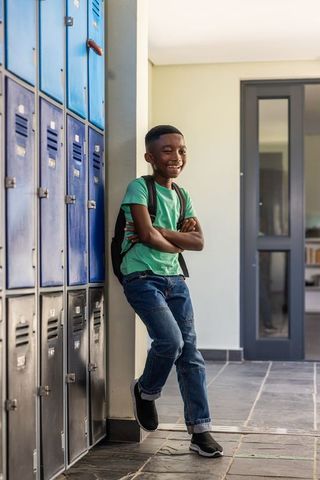 Smiling african american schoolboy leaning by lockers with backpack in bright hallway
