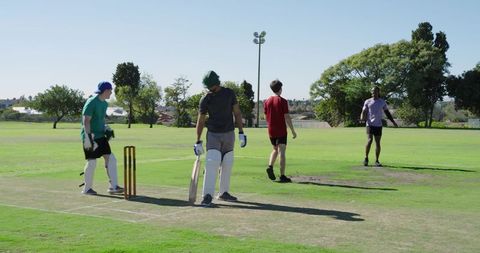 Batsman holding bat at crease during amateur cricket match on sunny park field with stumps
