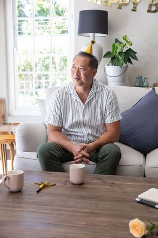 Senior asian man sitting on sofa wearing yellow party hat smiling during home birthday