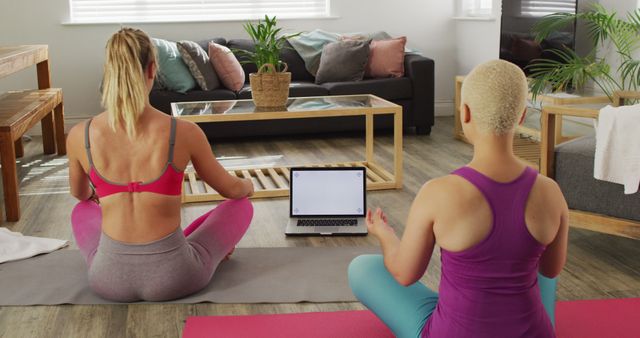 Two women are practicing yoga in a cozy living room using video guidance on a laptop. They are sitting on colorful yoga mats and focused on their meditation posture. Ideal for content promoting fitness routines, home workouts, wellness practices, and relaxing indoor activities.
