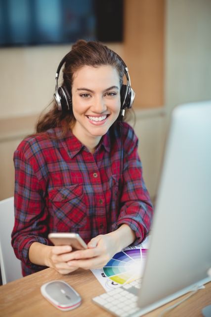 Young female graphic designer smiling while using mobile phone at her desk in a modern office. She is wearing headphones and a plaid shirt, indicating a casual and creative work environment. Ideal for use in articles or advertisements related to creative industries, modern workspaces, technology in design, or professional women in tech.