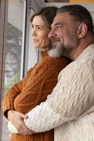Mature Couple Embracing by Window Wearing Cozy Cable-Knit Sweaters Showing Ring