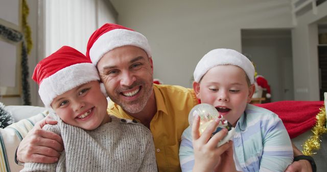 Father embracing two children in a cozy living room during Christmas season, showcasing festive joy and family bonding. Kids are wearing Santa hats and engaging with a snow globe, creating a warm holiday atmosphere. Ideal for use in advertisements, holiday cards, family-oriented marketing materials, and articles related to seasonal family activities.