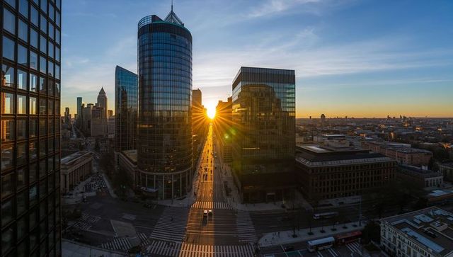 Sunburst aligning with downtown avenue framing cylindrical glass tower at urban sunset