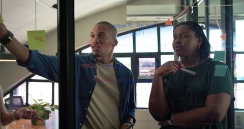 African american team brainstorming on glass wall with sticky notes in sunlit office