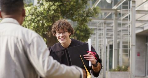 Smiling diverse male graduate greeting friend on campus holding diploma and mortarboard