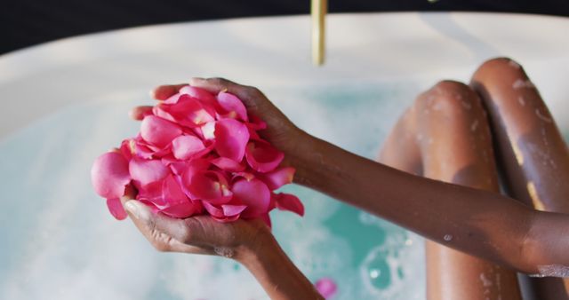 Person enjoying peaceful bath while holding pink rose petals in hands. Ideal for wellness, spa or self-care promotions, highlighting luxury and relaxation.