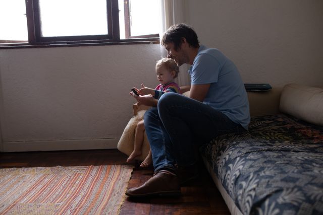 Side view of a young Caucasian father sitting on the sofa in a sitting room, with his baby, embracing him, holding smartphone in a hand, smiling.