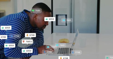 Mid Adult Man Leaning at Kitchen Counter Typing on Laptop with Croissant and Mug