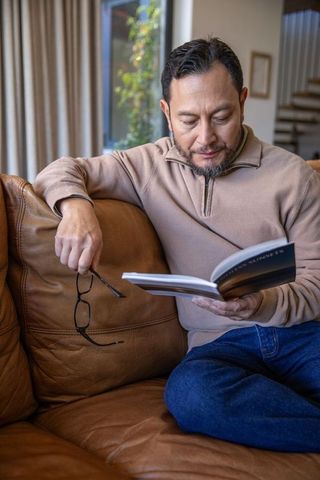 Mature asian man relaxing on leather sofa reading paperback book in cozy natural light