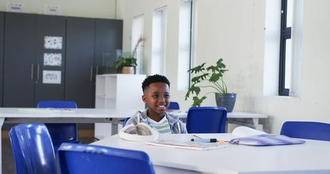 Smiling schoolboy sitting at classroom desk with headphones and colorful school supplies
