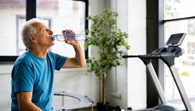 Senior man drinking water after workout in bright fitness center with treadmill