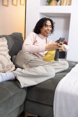 Non-binary person reclining on green sofa texting and smiling in cozy modern living room